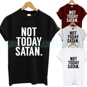 Puede incluir: Una colección de camisetas en negro, burdeos, blanco y gris claro. Cada camiseta presenta la frase "NOT TODAY SATAN" en letras mayúsculas blancas. Las camisetas se muestran en perchas de madera.
