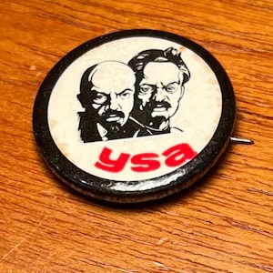 May include: A round black and white pin with two portraits and the red letters "ysa". The pin has a safety pin style clasp on the back. The portraits are of two men with serious expressions.
