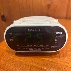 May include: A white Sony Dream Machine FM/AM clock radio. The digital display shows the time in green digits. The clock has alarm settings and radio frequency indicators. The clock is on a wooden surface.