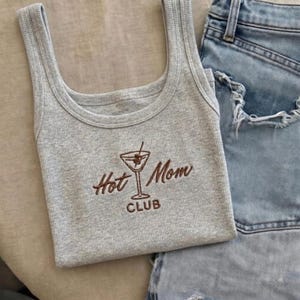 May include: A grey ribbed vest top featuring the embroidered phrase "Hot Mom Club" in brown, accompanied by a martini glass design. Light blue denim shorts are also visible.