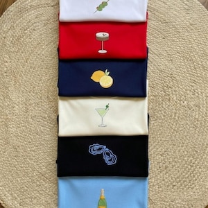 Puede incluir: Una pila de camisetas plegadas en varios colores, cada una con un bordado. Los diseños incluyen aceitunas, un cóctel, un limón, ostras y una botella de champán. Las camisetas están apiladas sobre una alfombra.