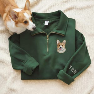 May include: A dark green sweatshirt with a quarter-zip collar and an embroidered corgi dog design. The sleeve cuff features the name "FINN" in white script. A real corgi dog is next to the sweatshirt.