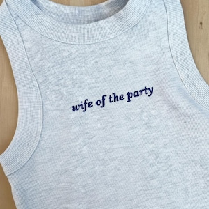 May include: A light gray ribbed tank top with the phrase "wife of the party" embroidered in navy blue. The sleeveless top has a rounded neckline and is made of a soft, comfortable material.