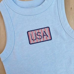 May include: A light blue ribbed sleeveless tank top featuring a rectangular embroidered patch on the chest. The patch contains the text USA in dark blue letters over a background of vertical red and white stripes with a dark blue border.