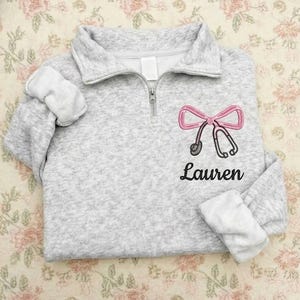 Könnte beinhalten: Hellgrauer Fleece-Pullover mit einem Viertel-Reißverschluss-Ausschnitt. Auf der Vorderseite befindet sich ein rosa Schleifen- und Stethoskop-Design, darunter der Name "Lauren" in schwarzer Schrift gestickt. Die Manschetten sind umgeschlagen und zeigen ein weißes Futter.