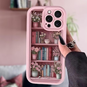 May include: Pink phone case featuring a bookshelf design with books, flowers, and a teacup. The case has a raised edge for camera protection and a smooth finish. The case is held in a hand.