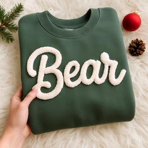 May include: A dark green sweatshirt with the word "Bear" in white, fluffy, textured letters. The sweatshirt is folded and held by a hand. A red bauble and pine cone are in the background.
