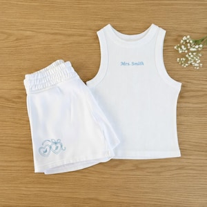 Puede incluir: Conjunto de camiseta sin mangas y shorts blancos acanalados. La camiseta sin mangas tiene el texto "Mrs. Smith" bordado en azul claro. Los shorts presentan un diseño de corazón azul claro. El conjunto se muestra sobre una superficie de madera.