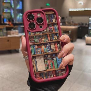 May include: A burgundy phone case featuring a detailed illustration of a bookshelf filled with colorful books, small decorative items, and a cup. The case has a raised edge around the camera lenses and a smooth, protective frame.