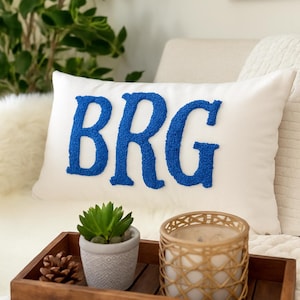 May include: A white rectangular pillow with the letters BRG embroidered in blue. A wooden tray holds a small potted succulent, a pinecone, and a candle in a woven holder. The pillow rests on a white surface.