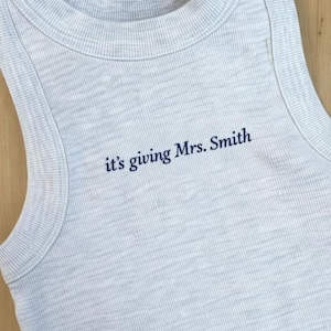 May include: Light gray ribbed tank top with the phrase "it's giving Mrs. Smith" in navy blue lettering. The sleeveless top has a rounded neckline and is made of a soft, textured fabric.