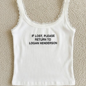 May include: White ribbed tank top with lace trim on the straps. The text "IF LOST, PLEASE RETURN TO LOGAN HENDERSON" is printed in black on the front of the top. The top is made of a soft, ribbed fabric.