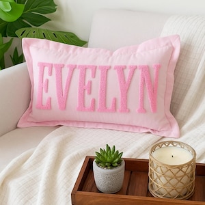 May include: A pink rectangular pillow with the name "EVELYN" embroidered in a textured, bubble-letter style. The pillow is on a white couch with a wooden tray holding a succulent and a candle.