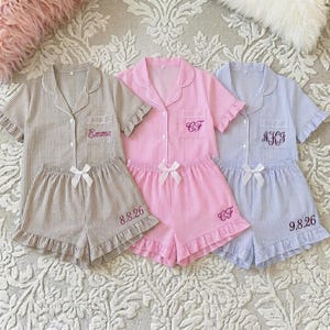 Op de afbeelding: Drie sets gestreepte pyjama's in bruin, roze en blauw. Elke set bevat een top met korte mouwen en een zak en een short met een strik. De bruine set heeft "Emma" geborduurd op de top en "8.8.26" op de short. De roze set heeft "CF" geborduurd op de top en short. De blauwe set heeft "AKG" en "9.8.26".