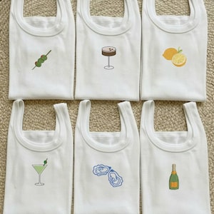 Puede incluir: Seis camisetas sin mangas blancas con diferentes diseños bordados de cócteles y alimentos. Los diseños incluyen aceitunas, un martini, un limón, ostras, una botella de champán y un cóctel.