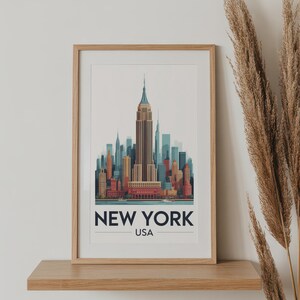 May include: A framed print featuring a stylized illustration of the New York City skyline, with the Empire State Building as the focal point. The print includes the text "NEW YORK USA" in a bold, modern font. The frame is made of light-colored wood.