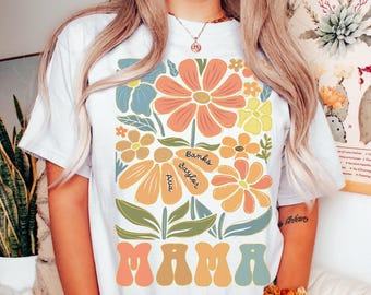 May include: White t-shirt with a floral design and the word "MAMA" in a retro font. The flowers have names written on them: Banks, Baylon, and Ava.