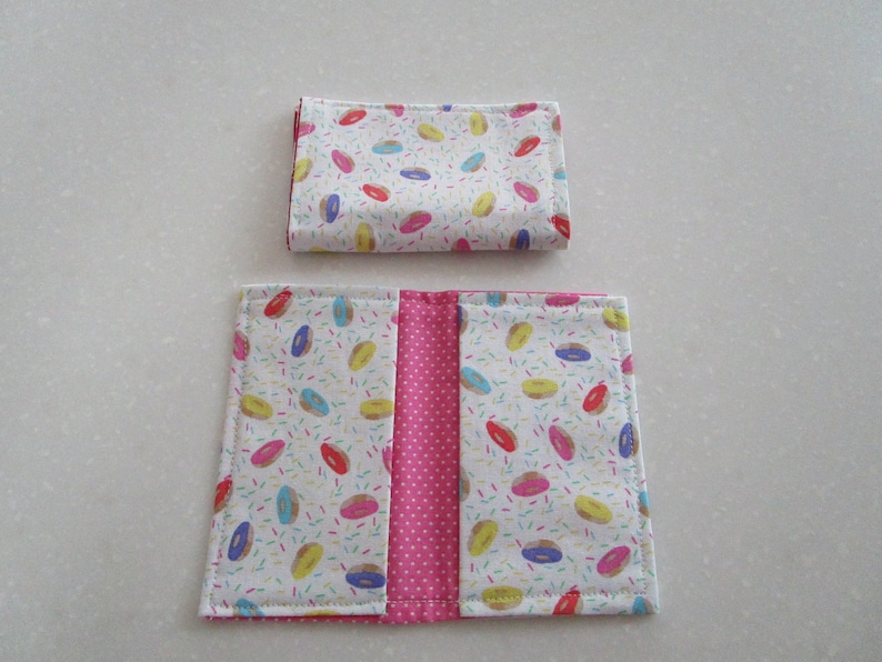 May include: A pink and white fabric wallet with a donut and sprinkles pattern. The wallet is folded in half and has a pink fabric lining.