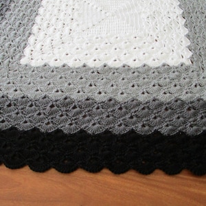 Pu&ograve; includere: Una copertina per neonati all'uncinetto con un quadrato centrale bianco, un quadrato esterno grigio e un bordo nero. La copertina ha un bordo a festoni.