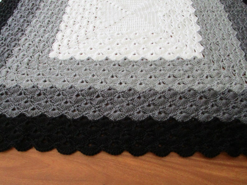 Pu&ograve; includere: Una copertina per neonati all'uncinetto con un quadrato centrale bianco, un quadrato esterno grigio e un bordo nero. La copertina ha un bordo a festoni.