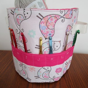 May include: A pink and white fabric storage bin with a bird pattern. The bin has a pink fabric lining with labeled compartments for organizing crochet hooks. A ball of gray yarn is inside the bin.