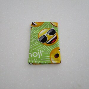 May include: A folded green fabric wallet featuring a repeating pattern of yellow emoji faces with sunglasses. The word "emoji" is printed in white on the wallet. The wallet is made of fabric.