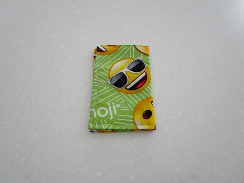 May include: A folded green fabric wallet featuring a repeating pattern of yellow emoji faces with sunglasses. The word "emoji" is printed in white on the wallet. The wallet is made of fabric.