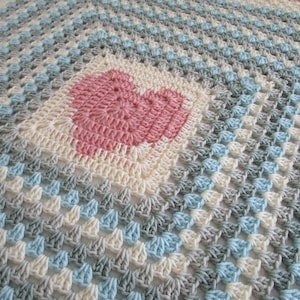 May include: A crocheted baby blanket with a pink heart in the center. The blanket is made with a variety of colors, including white, blue, and gray.