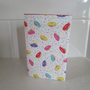 May include: A white fabric pouch with a colorful donut pattern. The donuts are pink, blue, yellow, and brown with white sprinkles. The pouch has a red zipper.