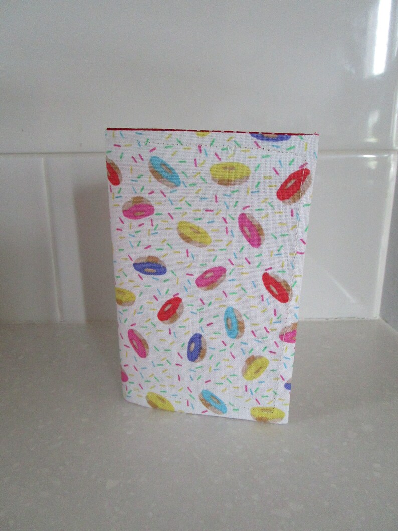 May include: A white fabric pouch with a colorful donut pattern. The donuts are pink, blue, yellow, and brown with white sprinkles. The pouch has a red zipper.