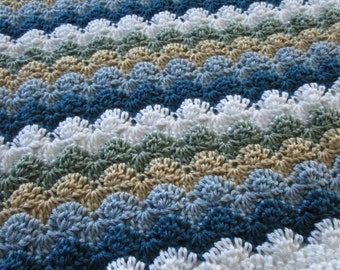 May include: A handmade crochet blanket with a wave pattern. The blanket features rows of varying colors, including white, shades of blue, green, and tan. The texture is visible, with each stitch creating a scalloped edge. The overall effect is a cozy, textured textile.