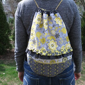 May include: A gray drawstring backpack with a yellow and white floral print. The backpack has a ruffled trim and a gray and white patterned bottom.