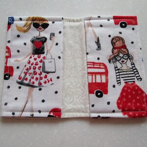 Puede incluir: Una cartera de tela blanca con un patr&oacute;n de lunares rojos y negros. La cartera presenta dos ilustraciones de mujeres con vestidos rojos y negros. Una mujer lleva una bolsa de compras y la otra lleva un par de gafas.