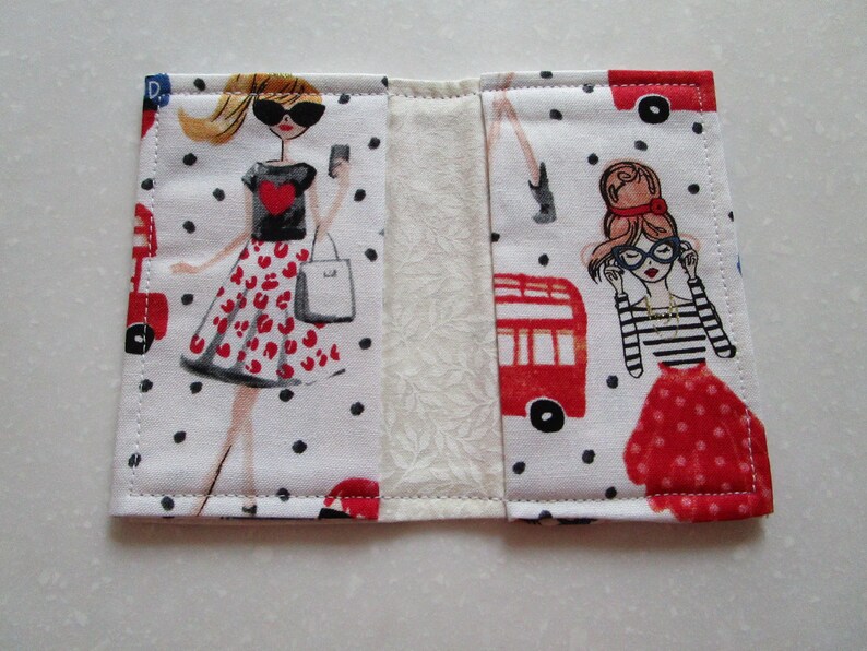 Puede incluir: Una cartera de tela blanca con un patr&oacute;n de lunares rojos y negros. La cartera presenta dos ilustraciones de mujeres con vestidos rojos y negros. Una mujer lleva una bolsa de compras y la otra lleva un par de gafas.