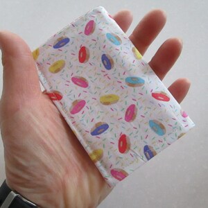 May include: A white fabric wallet with a donut and sprinkles pattern. The wallet is small and compact, perfect for carrying cards and cash.