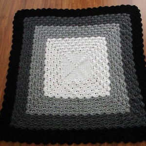 Pu&ograve; includere: Una copertina per neonati all'uncinetto con un quadrato centrale bianco circondato da un gradiente di quadrati grigi, il tutto incorniciato da un bordo nero.