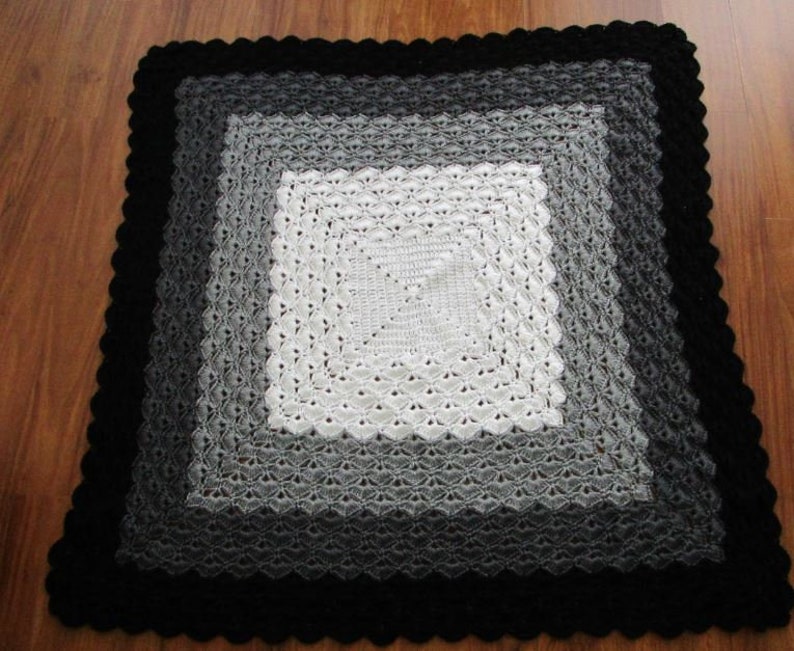 Pu&ograve; includere: Una copertina per neonati all'uncinetto con un quadrato centrale bianco circondato da un gradiente di quadrati grigi, il tutto incorniciato da un bordo nero.