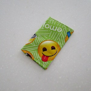 May include: A small, rectangular, green fabric item with a repeating pattern of yellow emoji faces and the word "emoji" in white. One emoji has its tongue sticking out. The item is likely a small pouch or case.