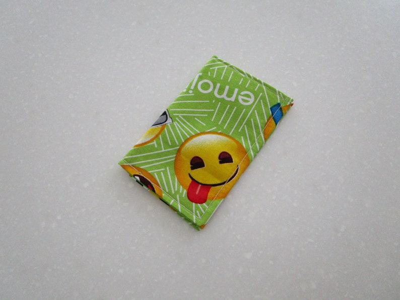 May include: A small, rectangular, green fabric item with a repeating pattern of yellow emoji faces and the word "emoji" in white. One emoji has its tongue sticking out. The item is likely a small pouch or case.