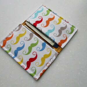 May include: A white fabric wallet with a colorful pattern of mustaches. The wallet is folded open to show the interior, which has a slot for cards and cash.