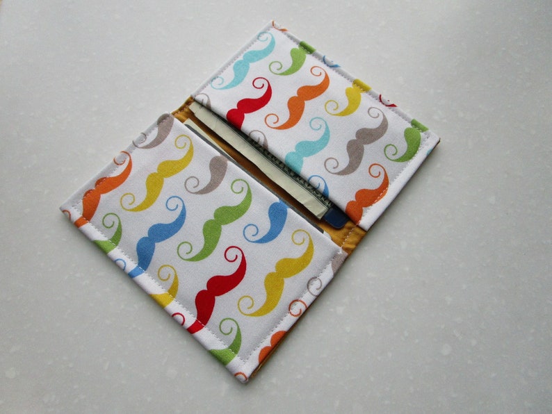 May include: A white fabric wallet with a colorful pattern of mustaches. The wallet is folded open to show the interior, which has a slot for cards and cash.