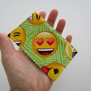 May include: A rectangular fabric item featuring a repeating pattern of yellow emoji faces with various expressions, including hearts for eyes and smiling faces. The background is light green with white lines. The item is held in a hand.