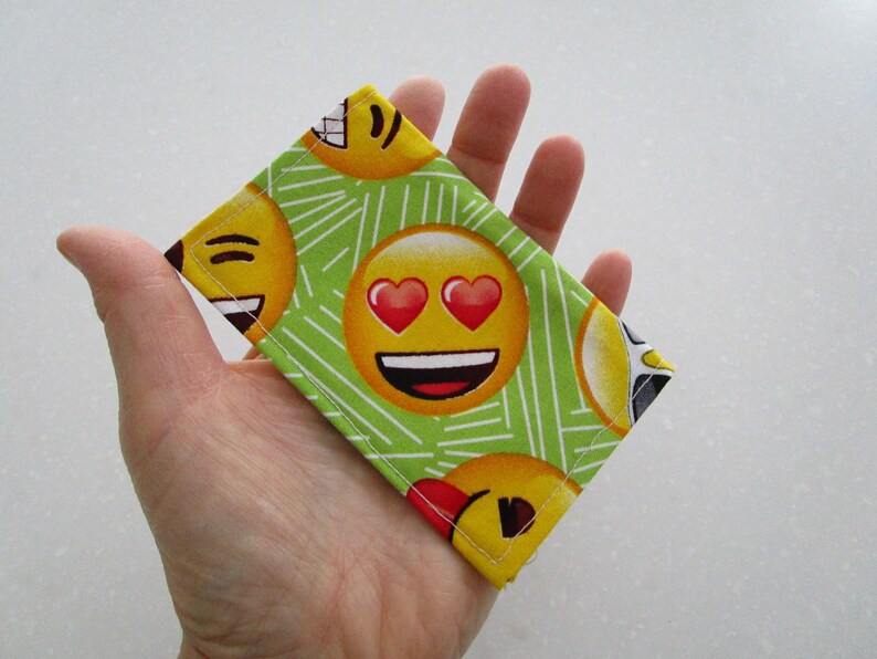 May include: A rectangular fabric item featuring a repeating pattern of yellow emoji faces with various expressions, including hearts for eyes and smiling faces. The background is light green with white lines. The item is held in a hand.