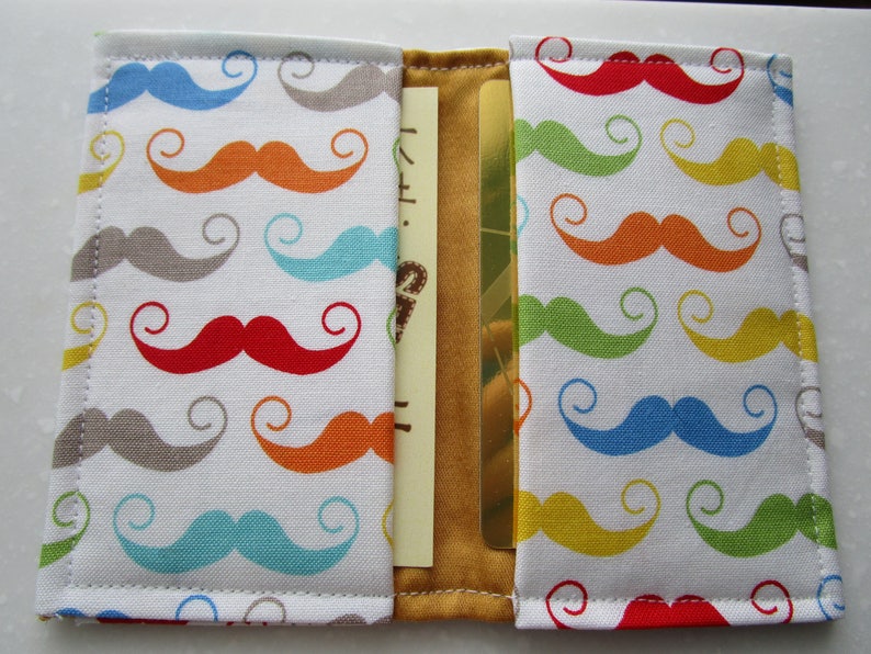 May include: A white fabric wallet with a gold card slot. The fabric has a repeating pattern of colorful mustaches with swirls.