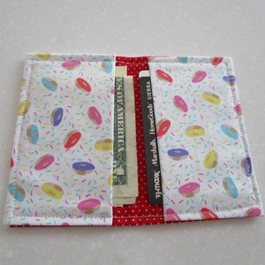 May include: A white fabric wallet with a donut and sprinkles pattern. The wallet is open and has a dollar bill and several credit cards inside.