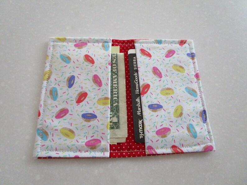 May include: A white fabric wallet with a donut and sprinkles pattern. The wallet is open and has a dollar bill and several credit cards inside.