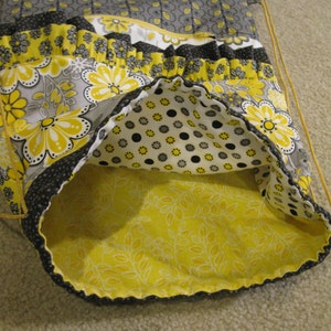May include: A yellow and gray fabric drawstring bag with a floral pattern and a black polka dot lining. The bag is made of cotton and has a ruffled trim.