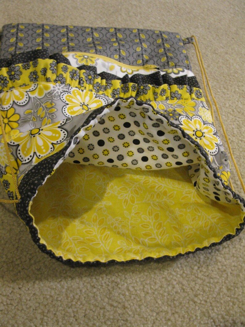 May include: A yellow and gray fabric drawstring bag with a floral pattern and a black polka dot lining. The bag is made of cotton and has a ruffled trim.