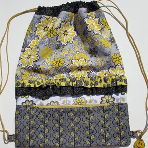 May include: A gray and yellow drawstring bag with a floral pattern and a zippered pocket. The bag has a black ruffle trim and a yellow smiley face zipper pull.