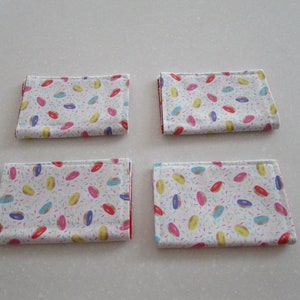 May include: Four white fabric napkins with a donut and sprinkles pattern. The donuts are pink, yellow, blue, and green.